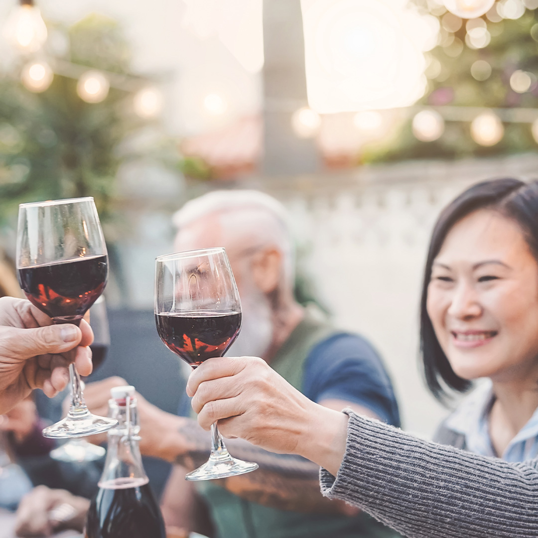 A group of diverse friends toasting with red wine outdoors, with warm string lights in the background.