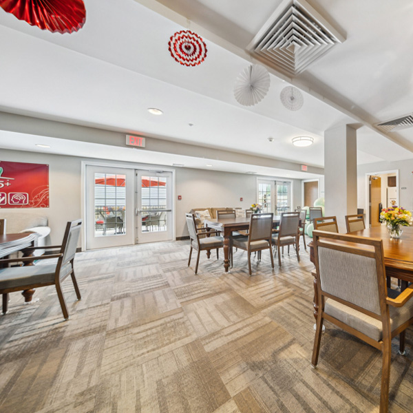 A bright community room with wooden dining tables, cushioned chairs, patterned carpet, and access to an outdoor patio.