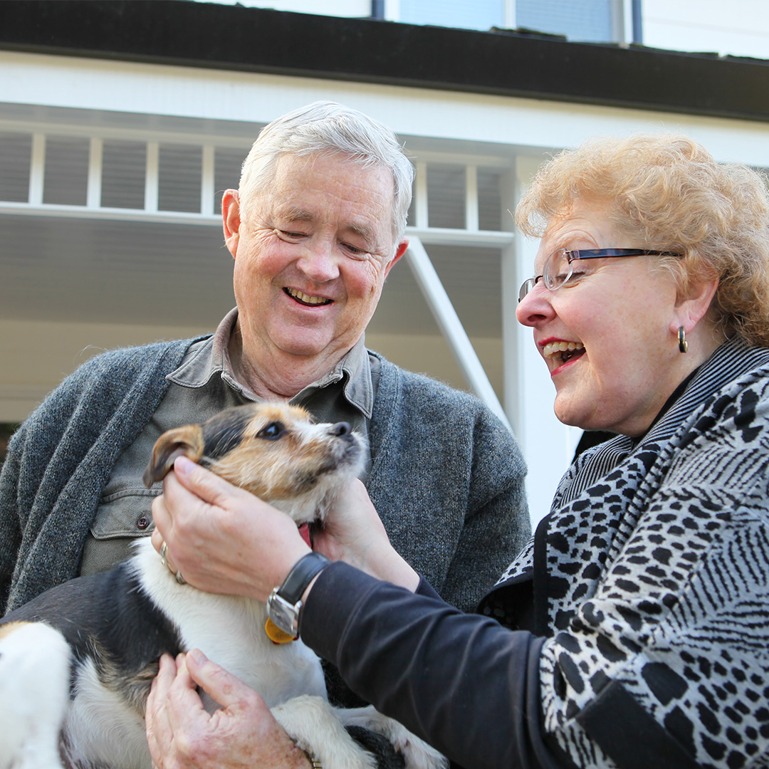 A laughing senior couple lovingly pets their small, happy terrier-mix dog outdoors in front of a white porch.