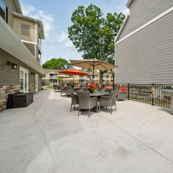 An exterior community patio featuring comfortable wicker chairs, dining tables with umbrellas, and a grilling area next to the buildings.