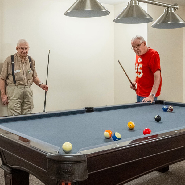 Two senior men playing a game of pool in a recreation room under metal hanging lights.