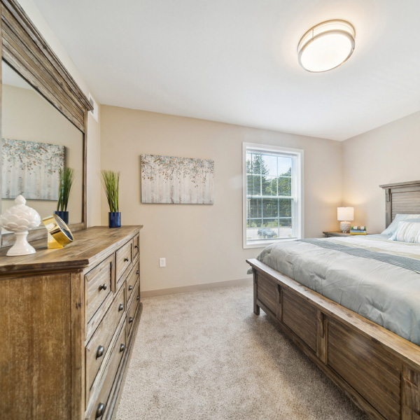 A bright bedroom featuring a large wooden bed, matching dresser, and neutral walls with carpet.