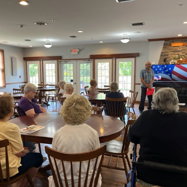 Man speaking to a small group of seniors in a community room with an American flag display on a screen.