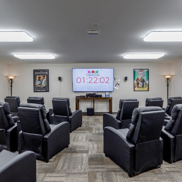 A dark community movie theater room with rows of black reclining chairs facing a large screen displaying "Showtime Starts In" countdown.