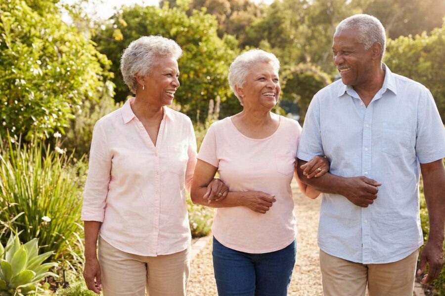 Three smiling older adults walk arm-in-arm on a garden path.