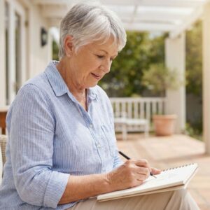 A senior person enjoying leisure time outdoors on a sunny patio, sketching in a notebook.
